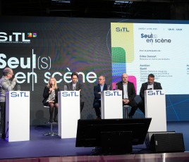Six personnes, cinq hommes, une femme, derrière quatre pupitres sur une scène lors d'une conférence. Un écran géant derrière eux.
