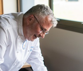 un chef cuisinier souriant, sur la gauche de la photo, penché sur une poêle, avec une fourchette et un couteau en main.