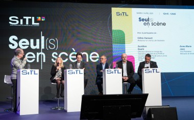 Six personnes, cinq hommes, une femme, derrière quatre pupitres sur une scène lors d'une conférence. Un écran géant derrière eux.
