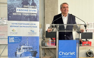 Au premier plan, un homme souriant fait un discours au pupitre ; une affiche au second plan. 