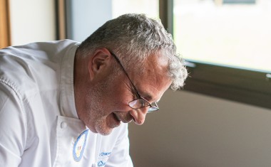 un chef cuisinier souriant, sur la gauche de la photo, penché sur une poêle, avec une fourchette et un couteau en main.