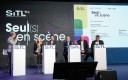 Six personnes, cinq hommes, une femme, derrière quatre pupitres sur une scène lors d'une conférence. Un écran géant derrière eux.