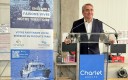 Au premier plan, un homme souriant fait un discours au pupitre ; une affiche au second plan. 