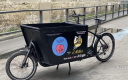 vélo cargo noir arrêté dans une rue. Logo bleu avec éléphant rose sur la caisse. 